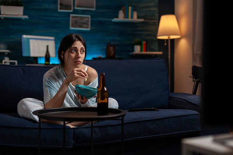A photo of a young woman looking sad watching tv alone drinking