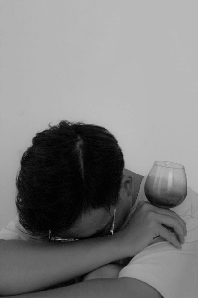 A photo of a man with his head between his arms holding a glass of wine