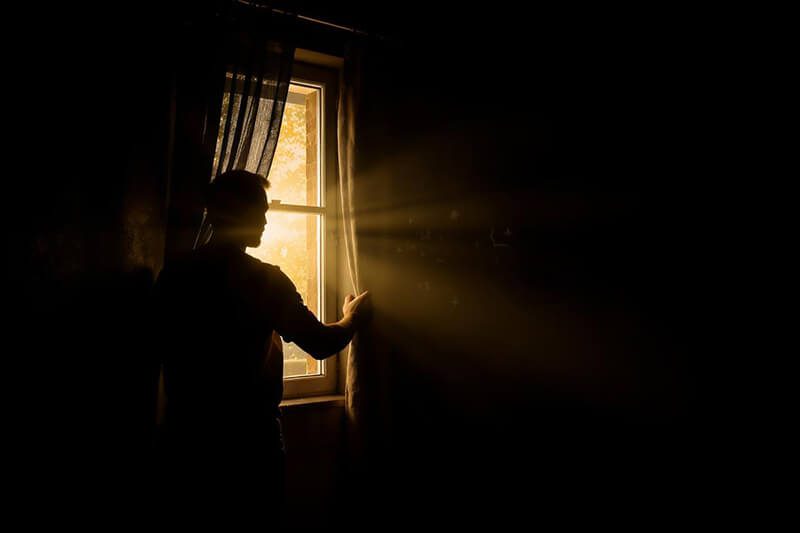 A photo of a man in a dark room pulling back the curtains and letting sunlight enter the room