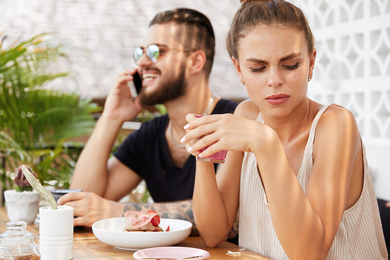 Photo of a couple at a restaurant, he is smiling while speaking on the phone, she looks uncomfortable