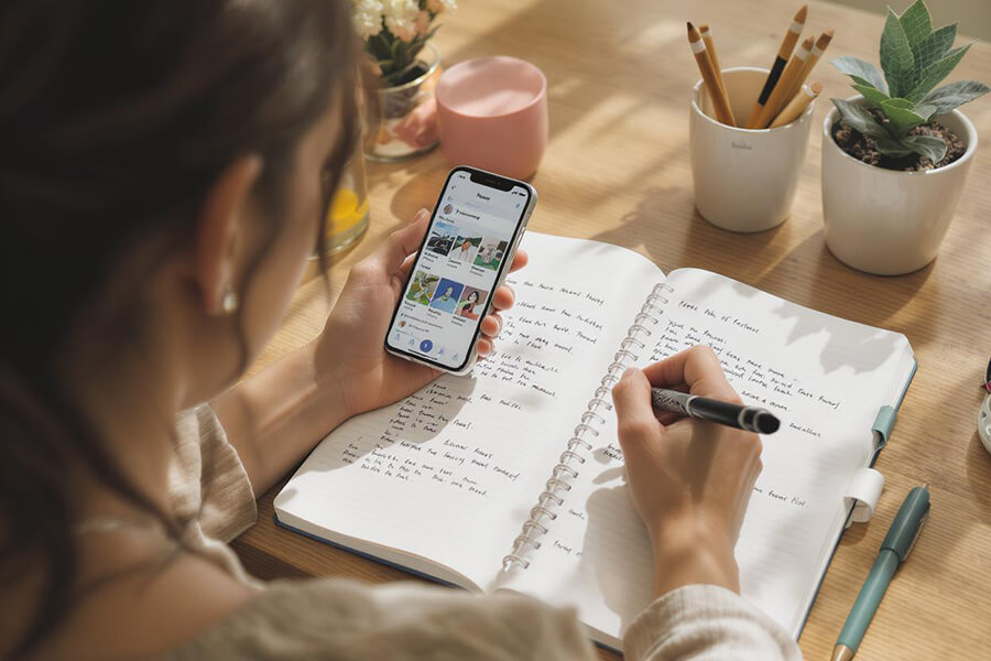 A photo of a woman writing in a journal and looking at social media on phone
