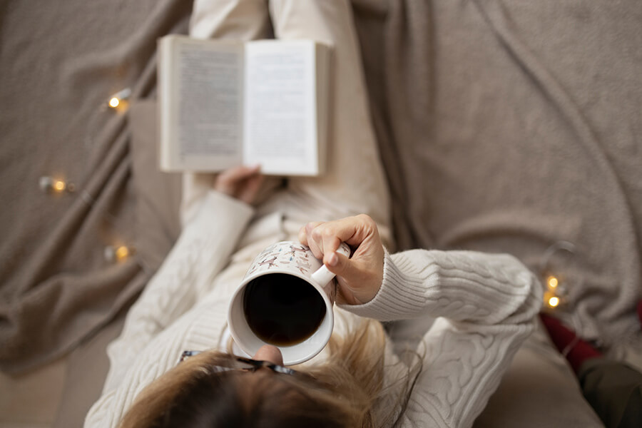 A photo of a woman drinking coffee while reading a book on the sofa