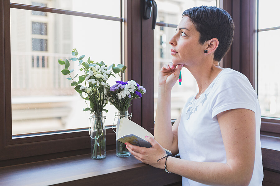 A photo of a woman looking out of a window holding a book